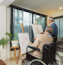 Elderly men ride a wheelchair doing hobby activity by painting in art school. Happy retired people having fun using creativity and practicing drawing at home.