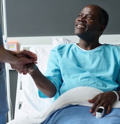 Elderly African American patient smiling to nurse while she holding his hands during her visit in ward